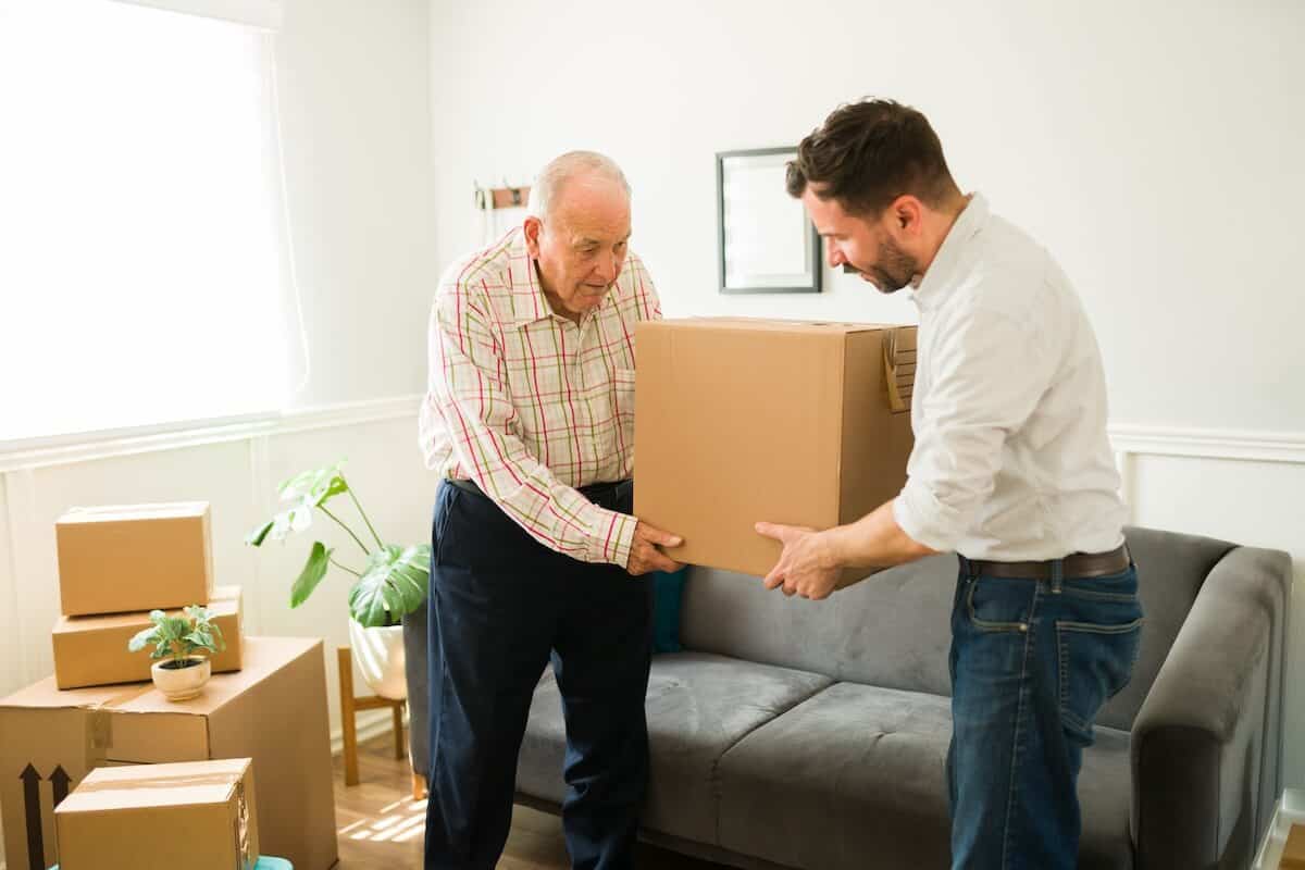 Move Into Senior Living-Young Man Carrying Boxes And Helping His Elderly Caucasian Father To Move To A New Apartment