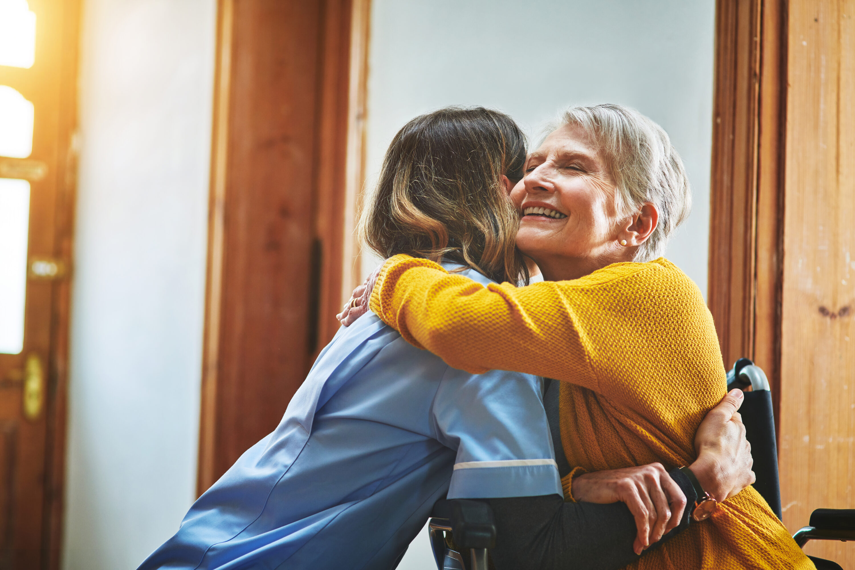 Elderly woman hugging caregiver in wheelchair indoors