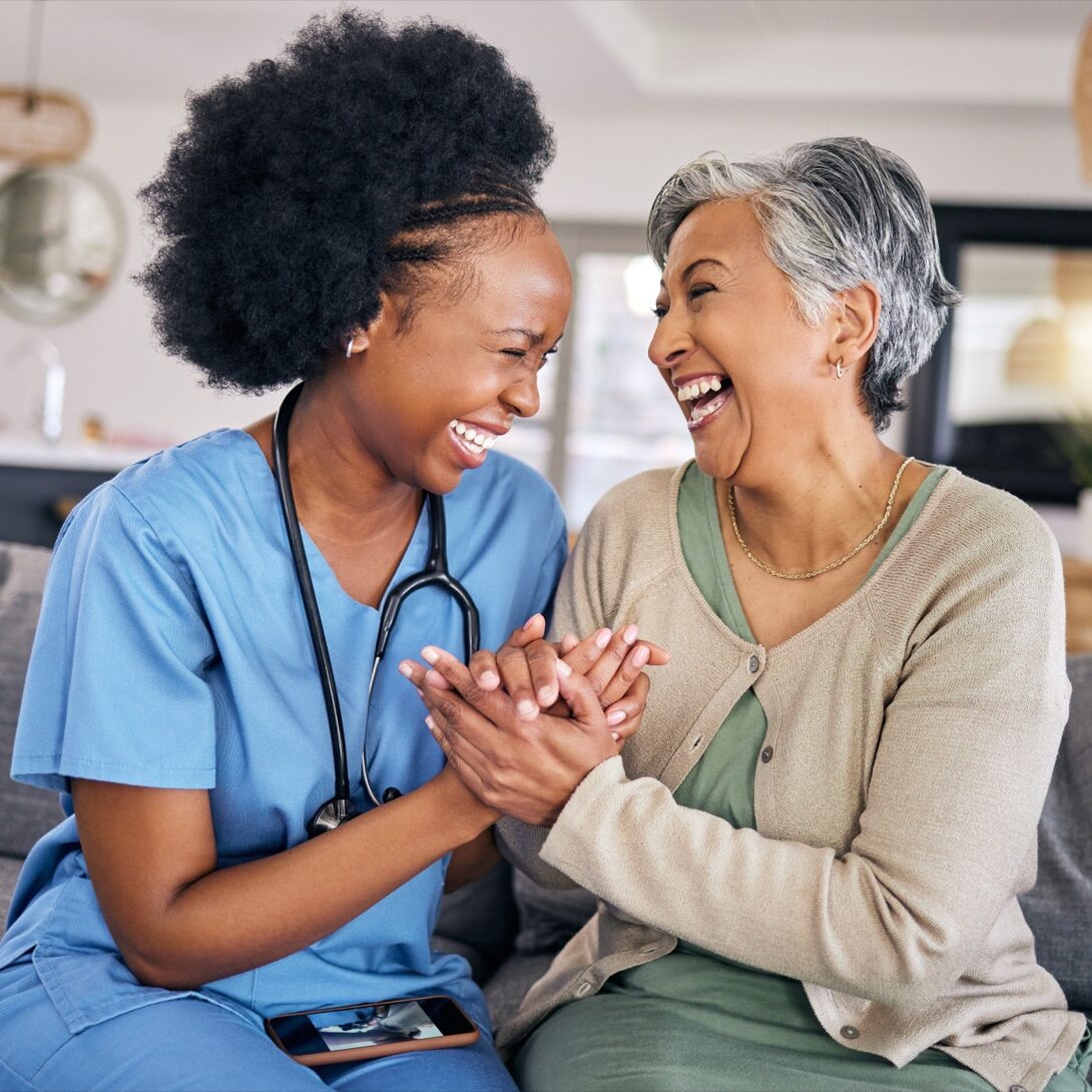 Nurse and older woman laughing, holding hands at home