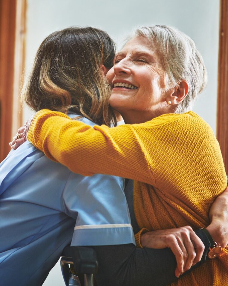 Elderly woman hugging caregiver in wheelchair indoors