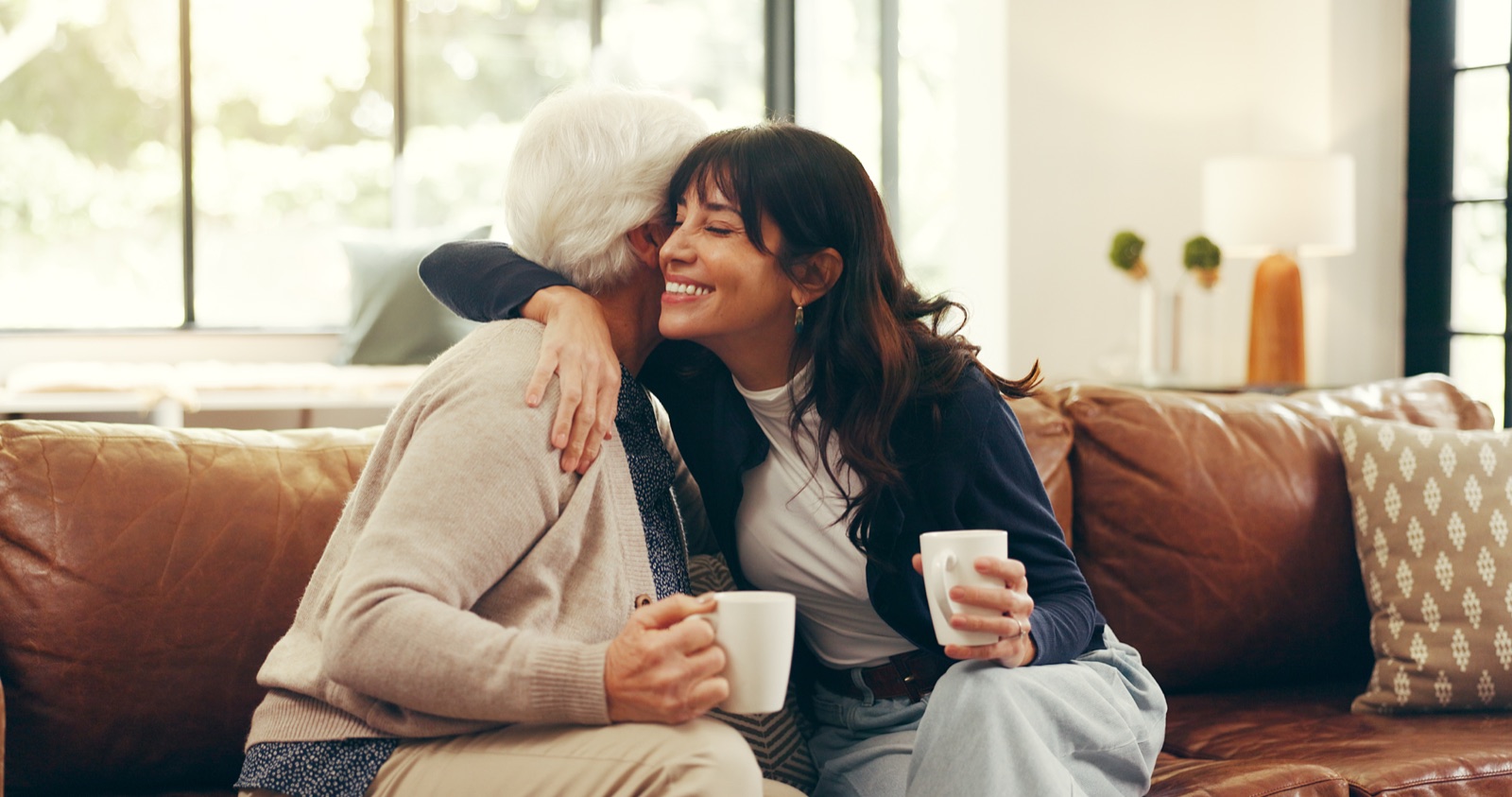 Daughter hugging elderly mother on couch with coffee